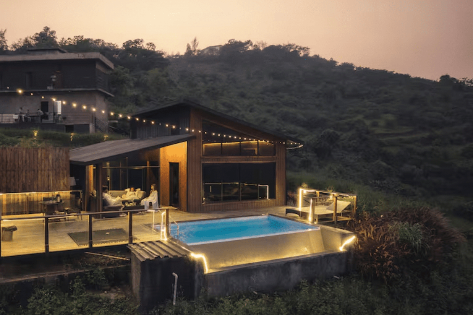 Illuminated villa with private pool and outdoor seating at dusk, surrounded by hills.