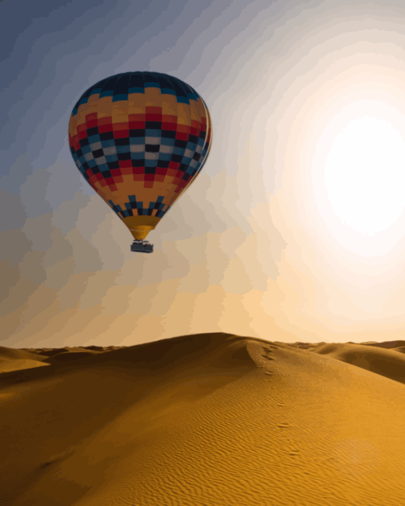 A colorful hot air balloon floats above the golden sand dunes under the morning sun in a desert landscape.