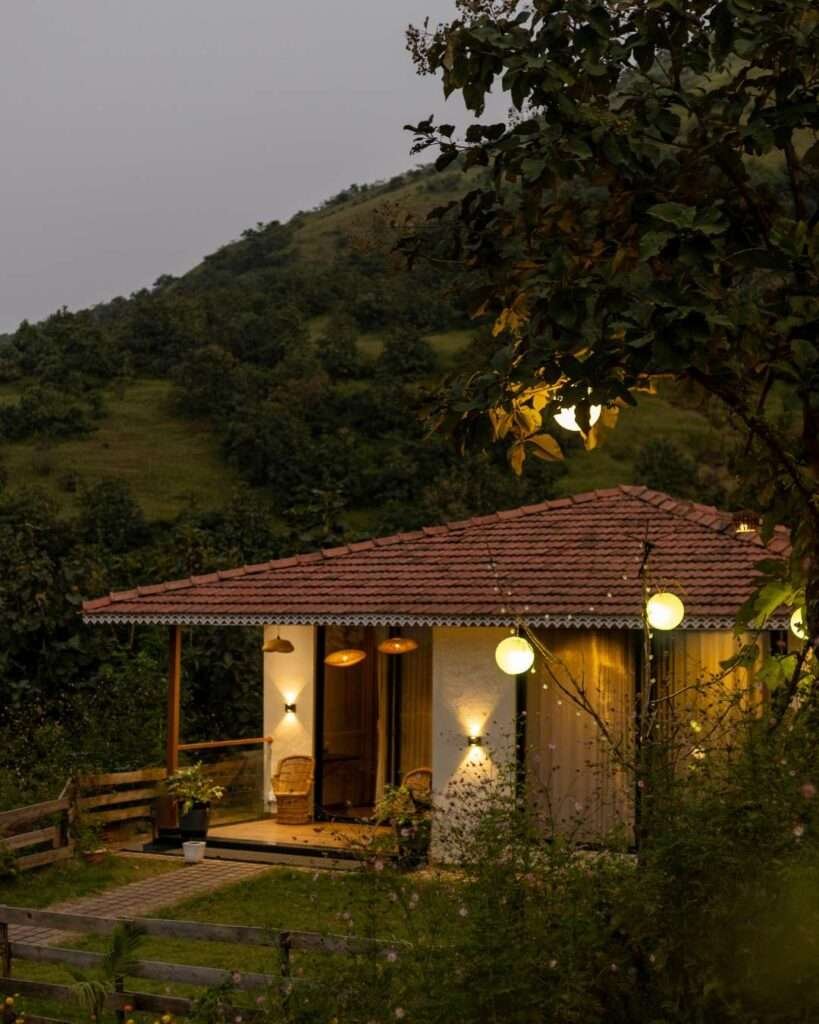 Cozy cottage with warm lights, set against green hills and surrounded by trees at dusk.