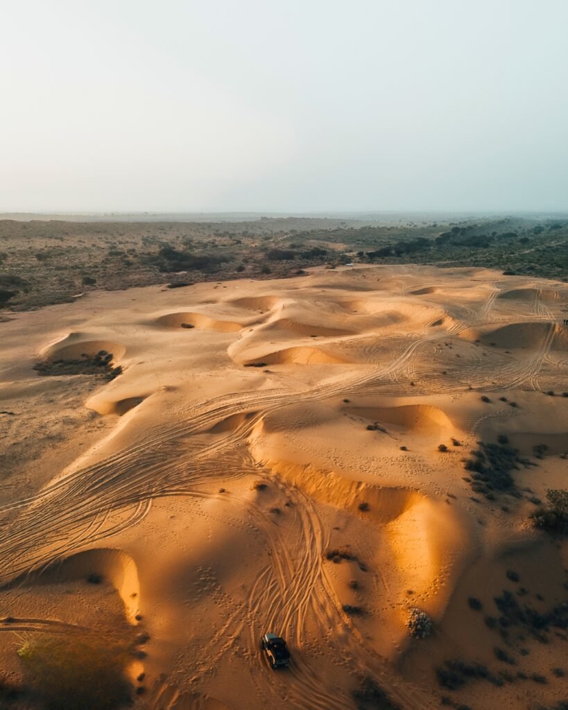 Aerial view of golden sand dunes in a desert with tire tracks and a vehicle.